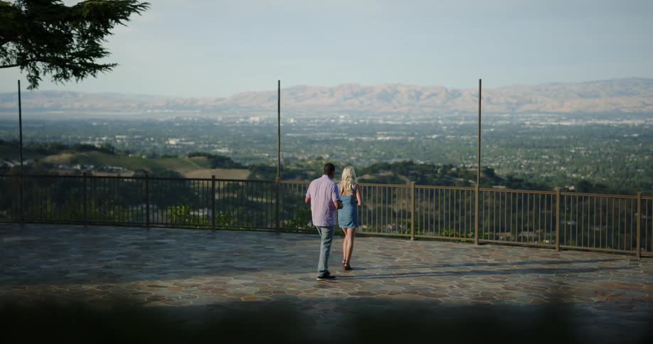 A couple walk to a railing with a view to drink wine