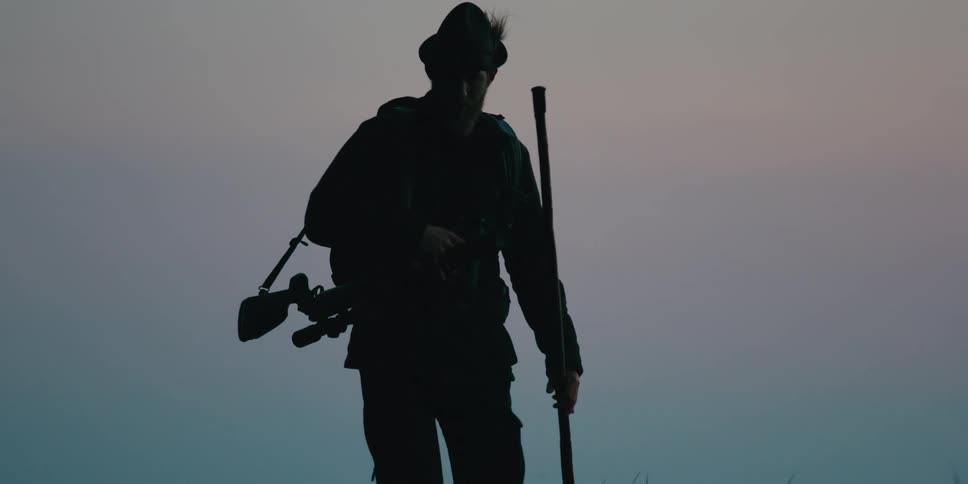 A man with a firearm slung over his shoulder looks through a set of binoculars before turning and leading a dog towards the camera