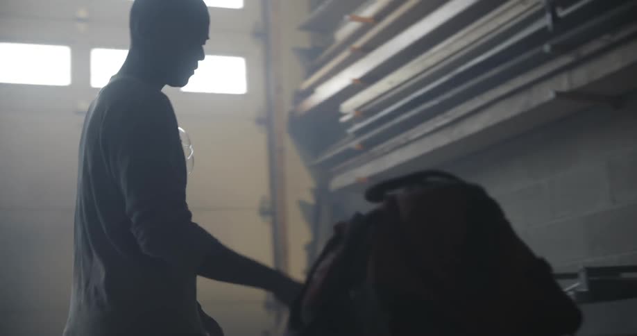 A man walks inside an industrial building and puts a bag down and picks up a piece of wood
