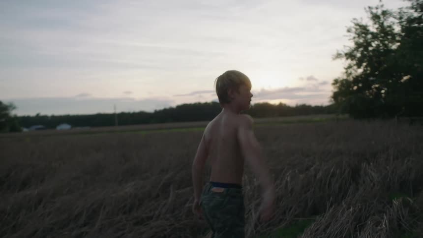 A young boy in a field with his father bends down and picks up his shirt and throws it