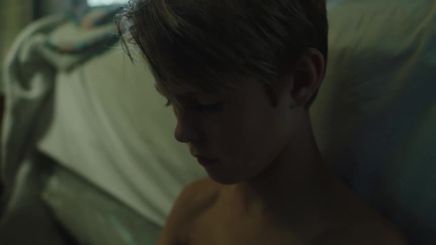 A shirtless boy sits on the floor of a room with his back resting against a bed