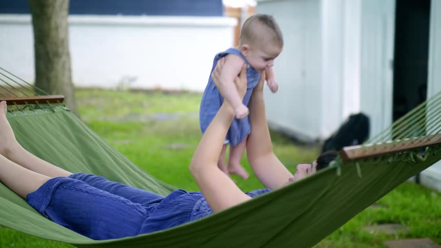 Woman lying in a backyard hammock hung between two trees holds her baby and moves it up and down in a playful way