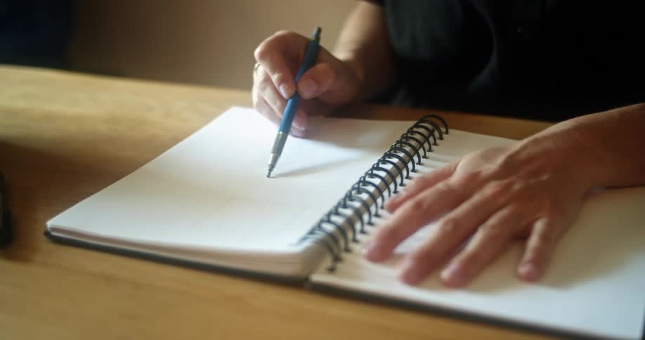 Female sitting at dining table holds open note book down with one hand while using a mechanical pencil to sketch on the other page