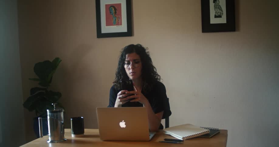 A woman typing on a laptop while sitting at a table stops to check her phone