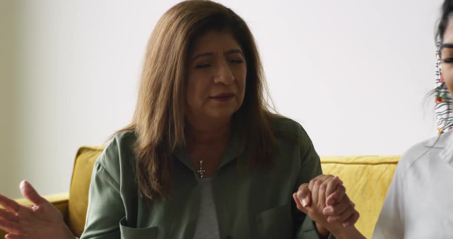 A woman sits on the couch with another woman and holds her hand while praying