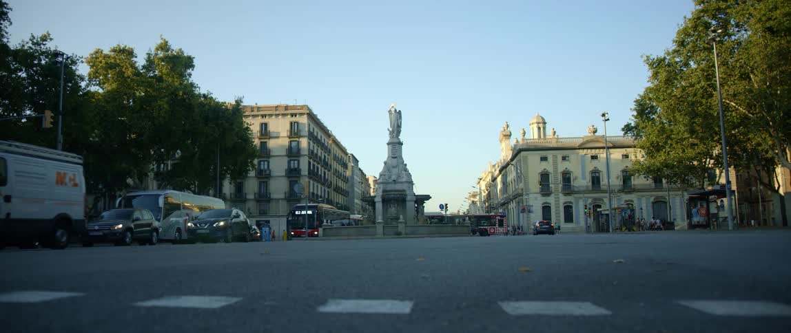 City buses and personal vehicles drive across a crosswalk in a city near a monument