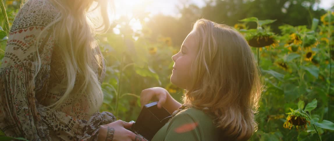 A girl holds a large old fashioned camera as she stands in a field of tall flowers with her mother during the day