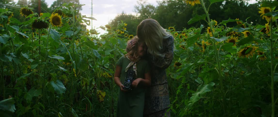 A girl holding a camera while standing in a field of tall flowers has her hair stroked and forehead kissed by her mother