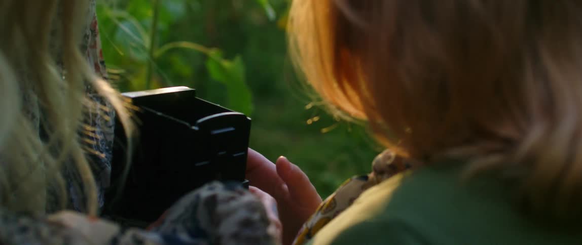 A woman and a girl holding and looking at a camera with flowers and trees in the background
