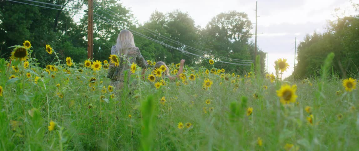 A woman holds hands with a girl as they walk through a field of tall grass and flowers