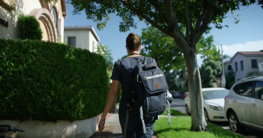A woman wearing a backpack walks along a sidewalk through a residential neighborhood on a sunny day