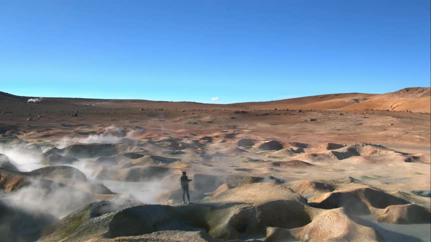 A man walks across terrain covered with geysers and takes pictures