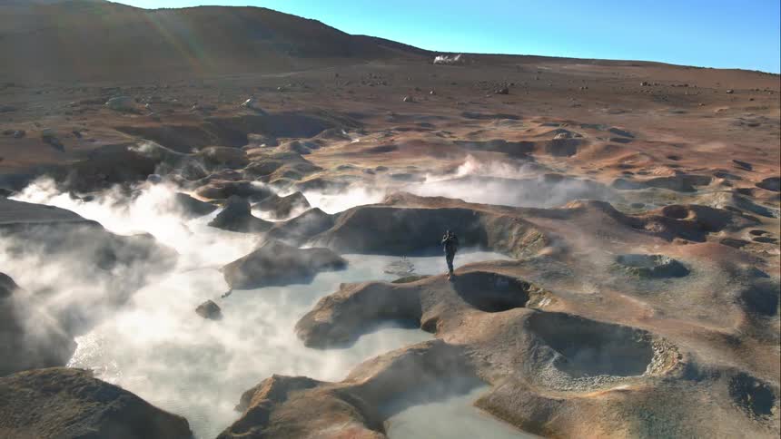 A photographer walks along a barren field of geysers before stopping to take a photograph