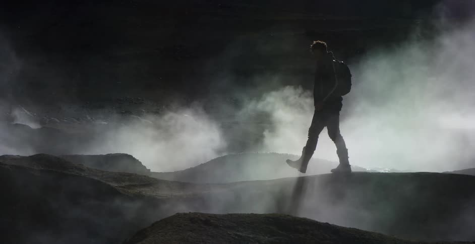 A photographer walking through a dark landscape riddled with geyser at night raises his camera to take a photo