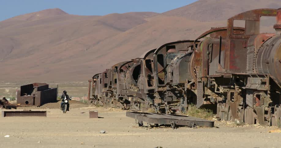 Man with a black leather jacket rides a motorcycle next to a train wreck in the desert