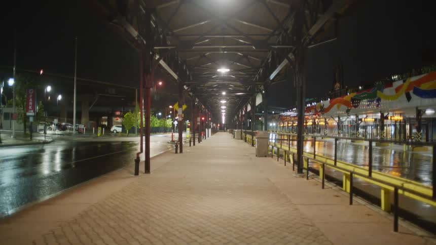 Brightly lit covered walkway separates wet street from parking lot near stores at night