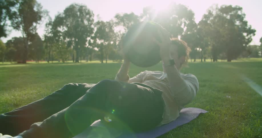 A man holds an exercise ball and leans backwards on a yoga mat and then up again while in a public park