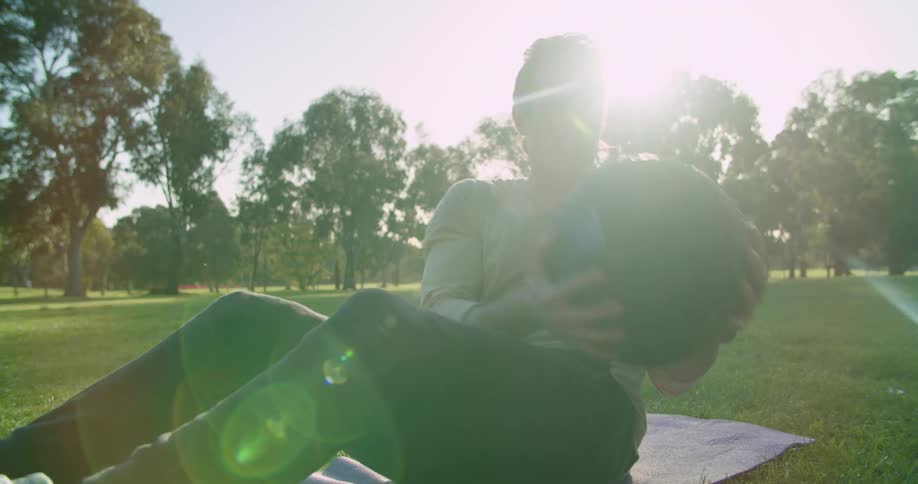 Man exercises with ball sitting on blanket in lawn in park on sunny day