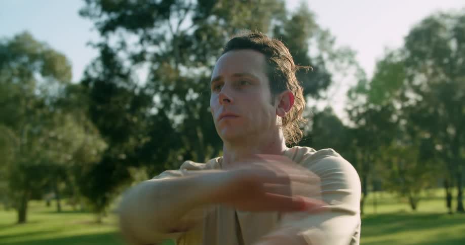 A white man stands in a park stretching his arms