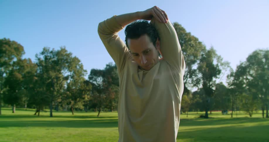 Man with long hair and a brown sweatshirt warms up before working out on green grass in the public park
