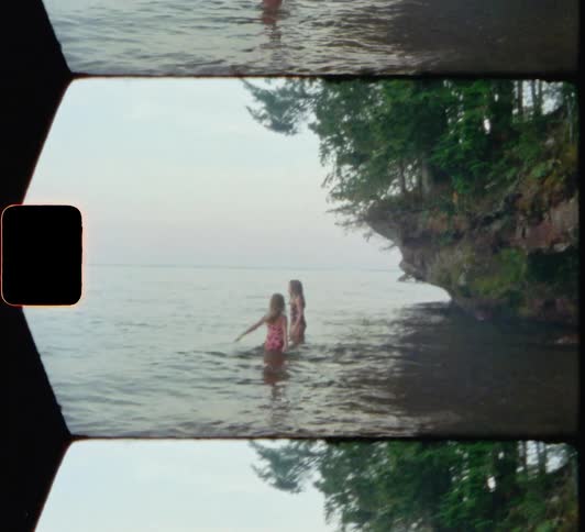 Two blonde girls wade and play in ocean near shore and tree covered promontory