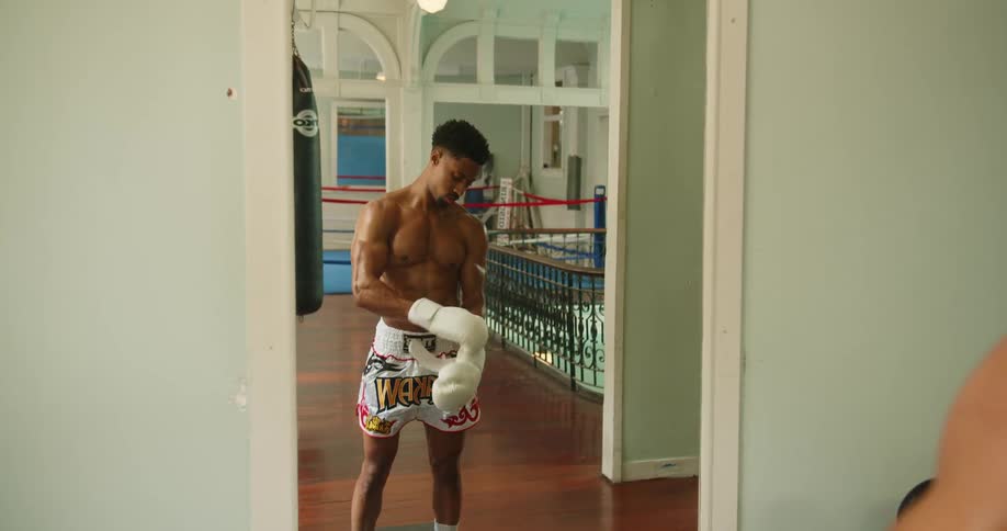 Muscular boxer with white boxing shorts wears white boxing gloves in front of a mirror in the gym