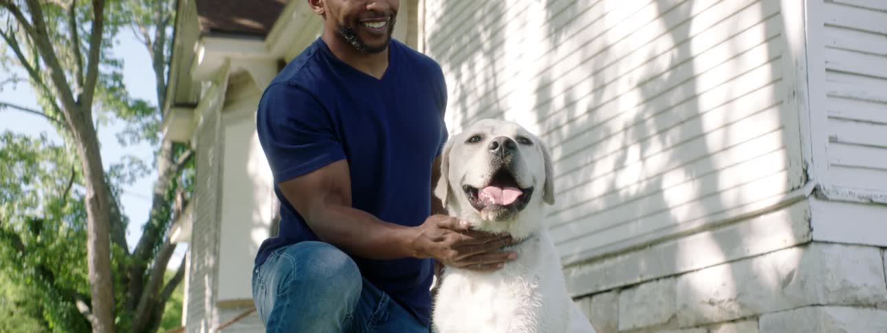 A black man smiles while petting a white dog outside of a house