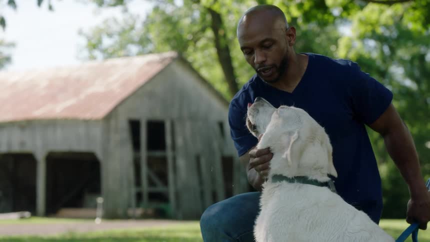 A man kneels down and pets his dog outside with a worn barn in the distance