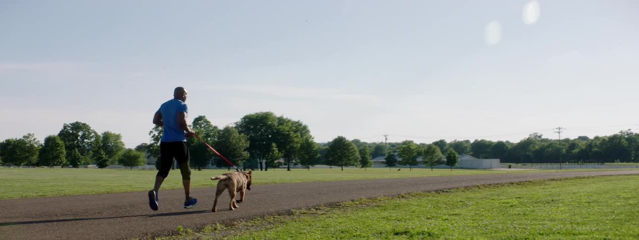Athletic man with a pet dog on a leash jogging on a paved road on a park