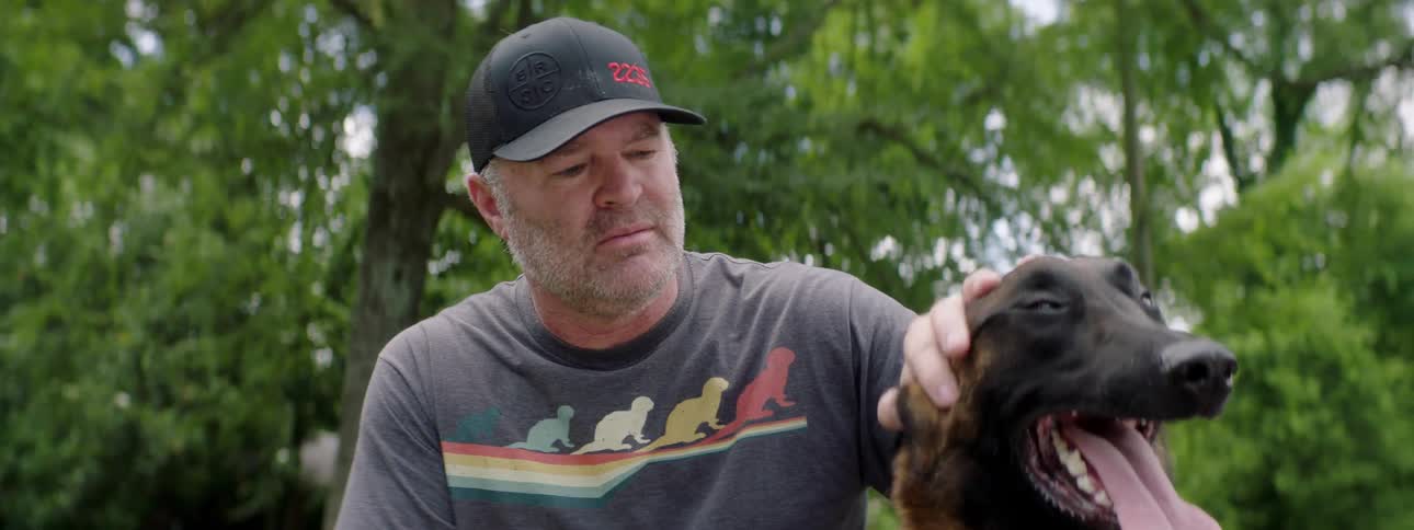 Middle aged man with a cap and a beard sitting on the sidewalk pets a brown dog