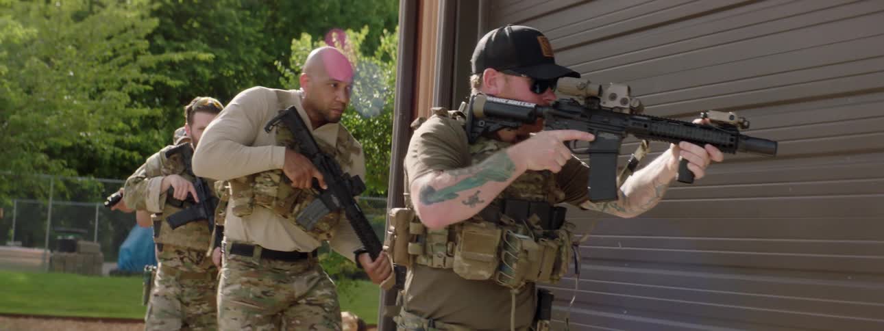 A group of armed soldiers walk around the corner of a building and approach its door