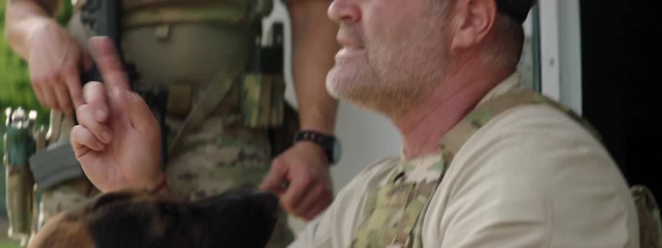 Man sits and pets german shepherd while talking to other men carrying weapons