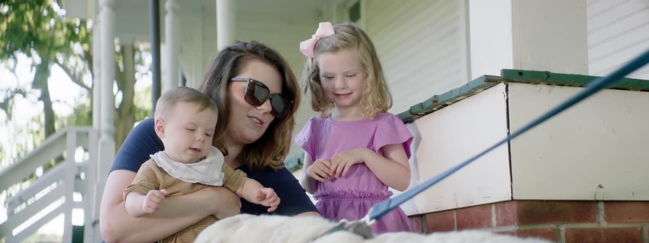 Mother crouches down holding baby near daughter as she pets white dog on leash