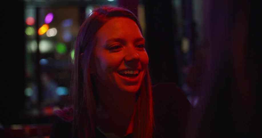 A young woman laughs while sitting at a table with other women in a bar at night