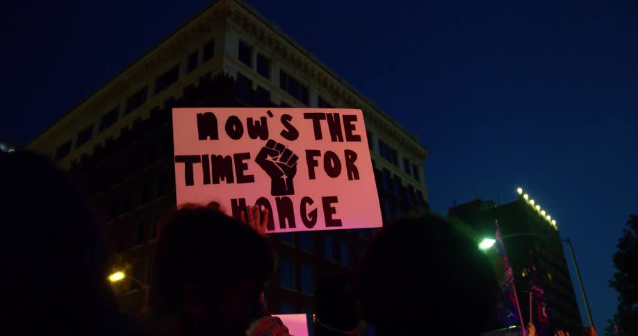 People protesting holding up signs with buildings trees and the sky in the background