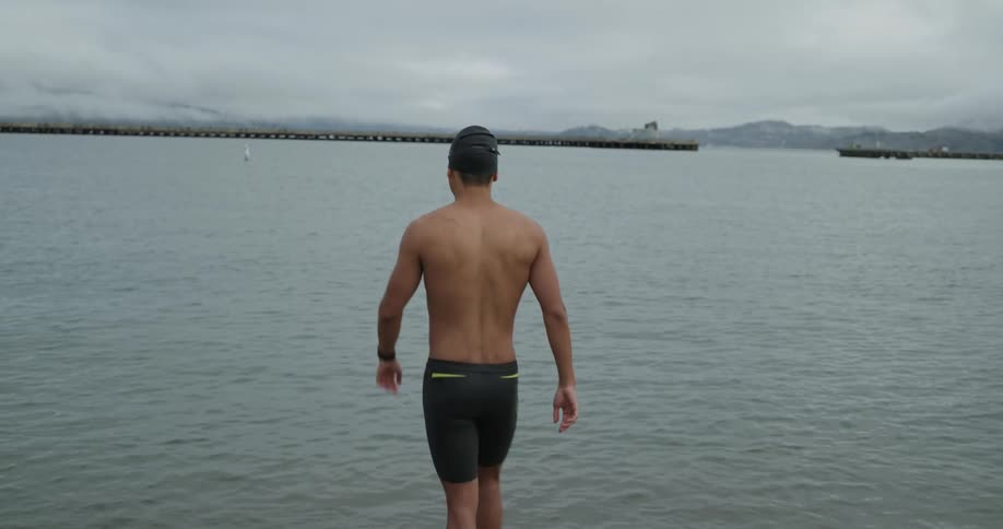 A man in a swimming cap and trunks sets his watch before slowly walking into the ocean on a cloudy day