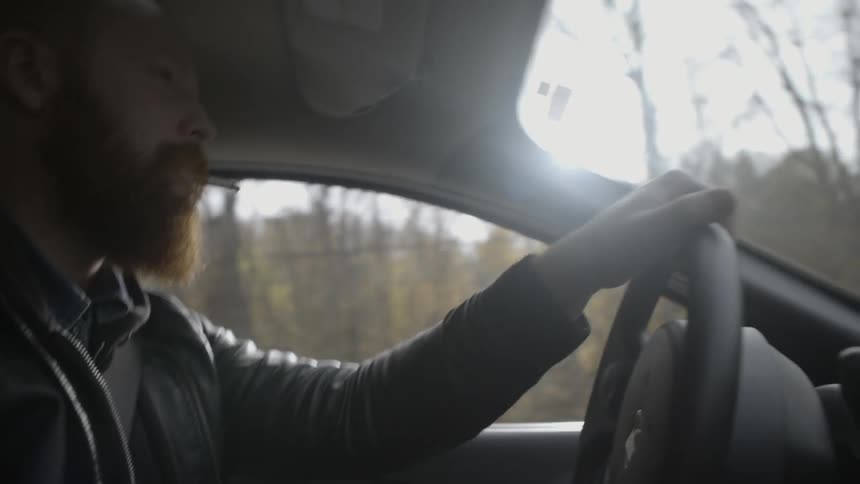 A bearded man smokes a cigarette while driving a car