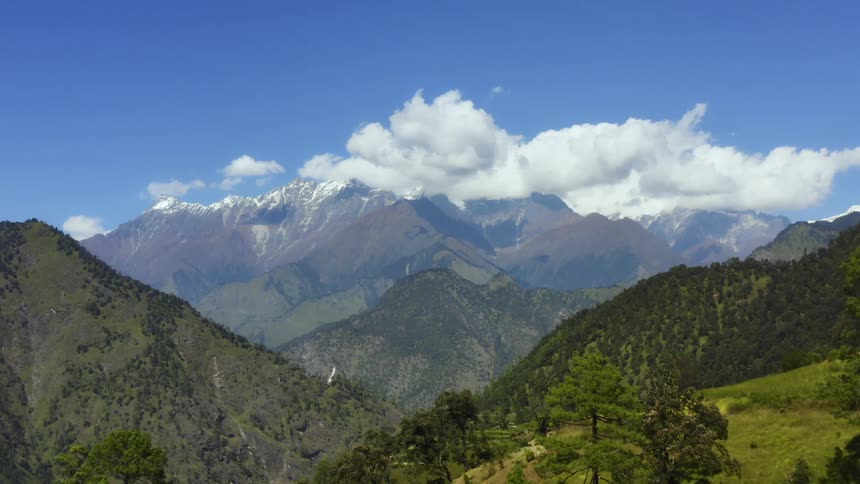 Clouds loom over a snow capped mountain standing over a series of smaller mountains covered in vegetation and forest