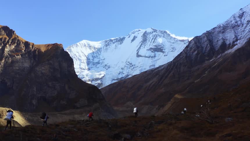 A line of hikers carry thick backpacks as they hike through a mountain valley on a sunny day