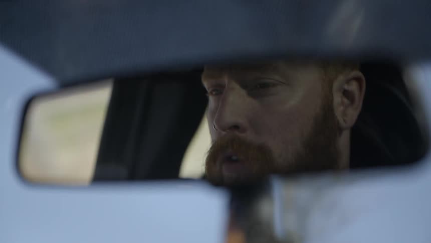A man's reflection is shown in the rearview mirror while he drives a car