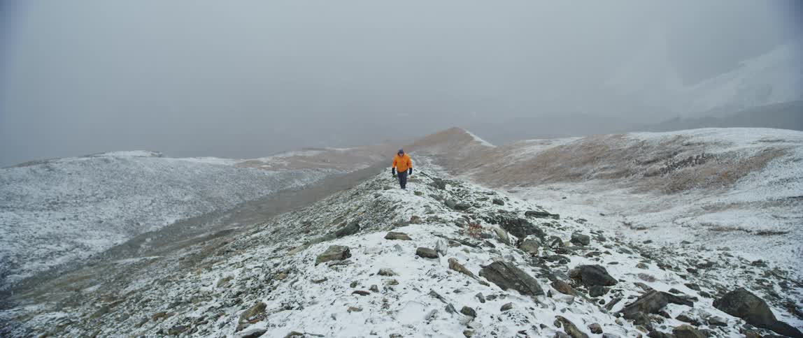 A man walks along a snowy rock strewn hill as it snows on a foggy day