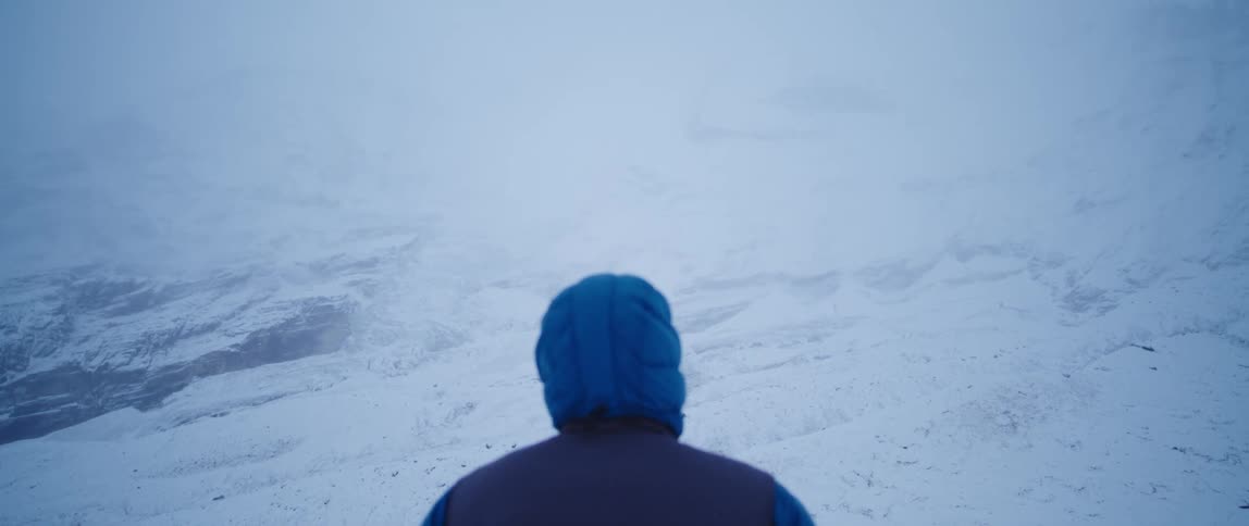 A man in a blue puffer coat stands with his hands in his pockets on a snow covered slope