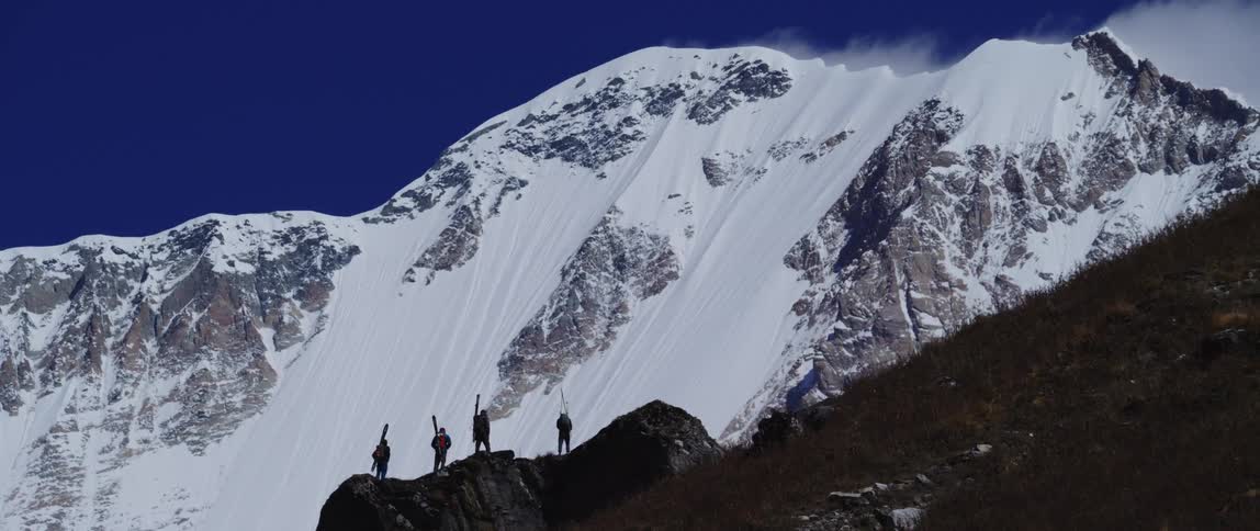Four people holding skis stand and look up a mountain