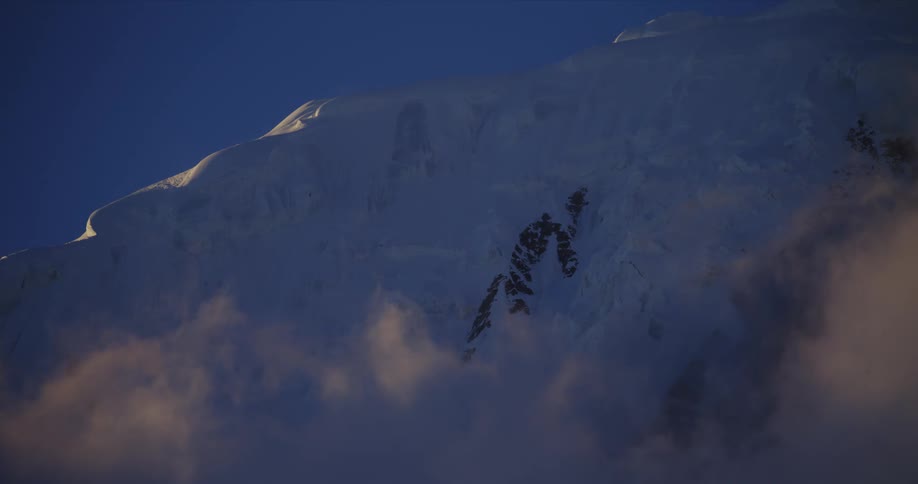 A large mountain ridge is almost completely covered in snow as clouds drift nearby it