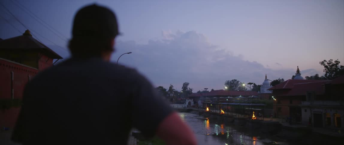 A man leisurely standing outdoors in a city in the evening looks across a small river towards a row of campfires on the opposite shore