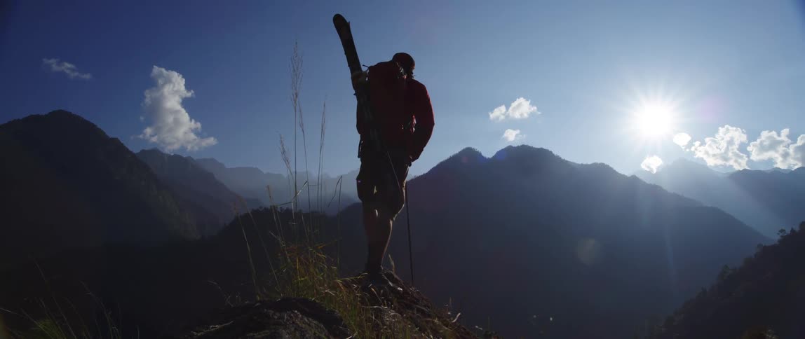 Man carrying skis in backpack stands on peak looking out on mountains under bright sunny sky