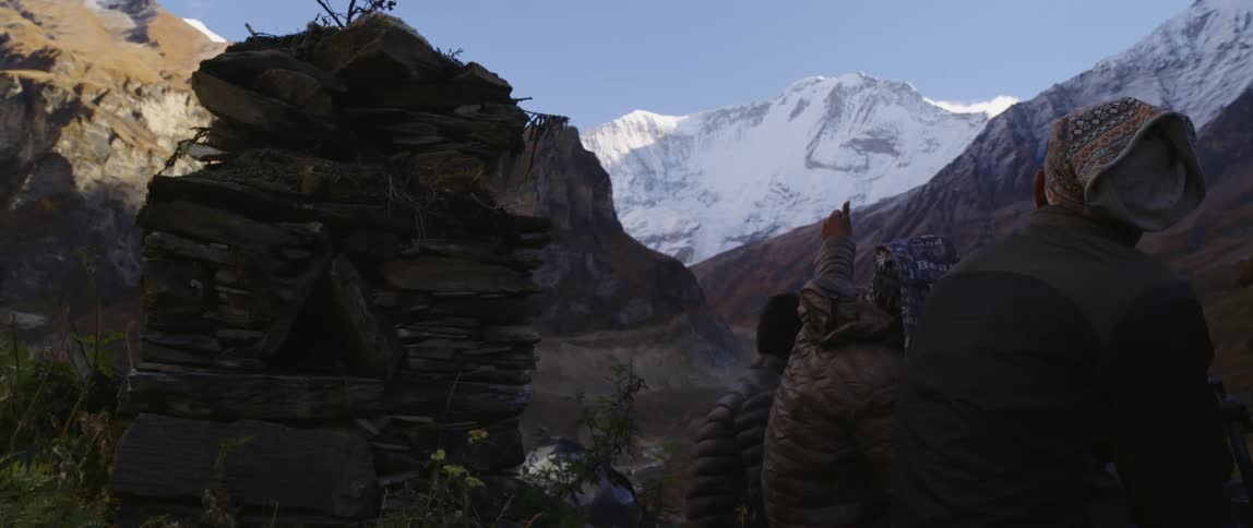 People sit in shadowy valley near small tower of stacked stones looking and pointing toward snow covered mountain