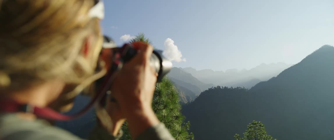 A blonde photographer takes a photograph of a mountain peak in the distance