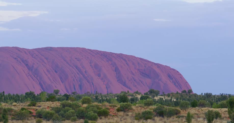 Uluru Ayers Rock looms over shrubland on a sunny day