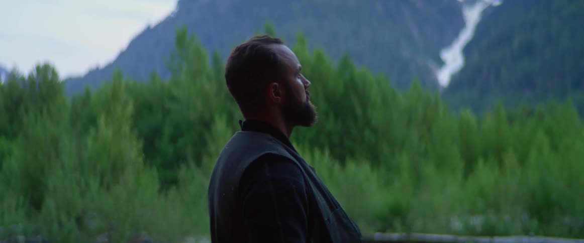 A man wearing a suit while standing in the wilderness looks up at a mountain covered in vegetation